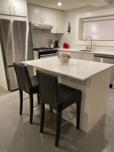 A modern kitchen is presented with sleek white cabinetry and stainless steel appliances. A minimalist island features a white countertop, complemented by two black woven bar stools. Natural light enters through a nearby window, enhancing the neutral tones of the space.