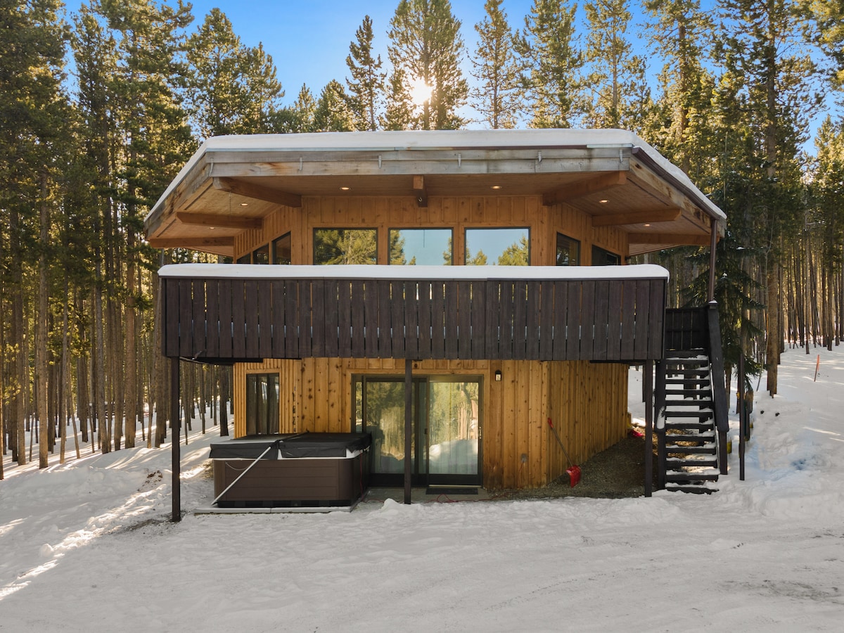 A modern hexagon-shaped home is nestled among tall trees, showcasing a wooden exterior and large windows. Snow blankets the ground, and a private hot tub is positioned on the lower deck. A staircase leads to the upper level, enhancing accessibility.
