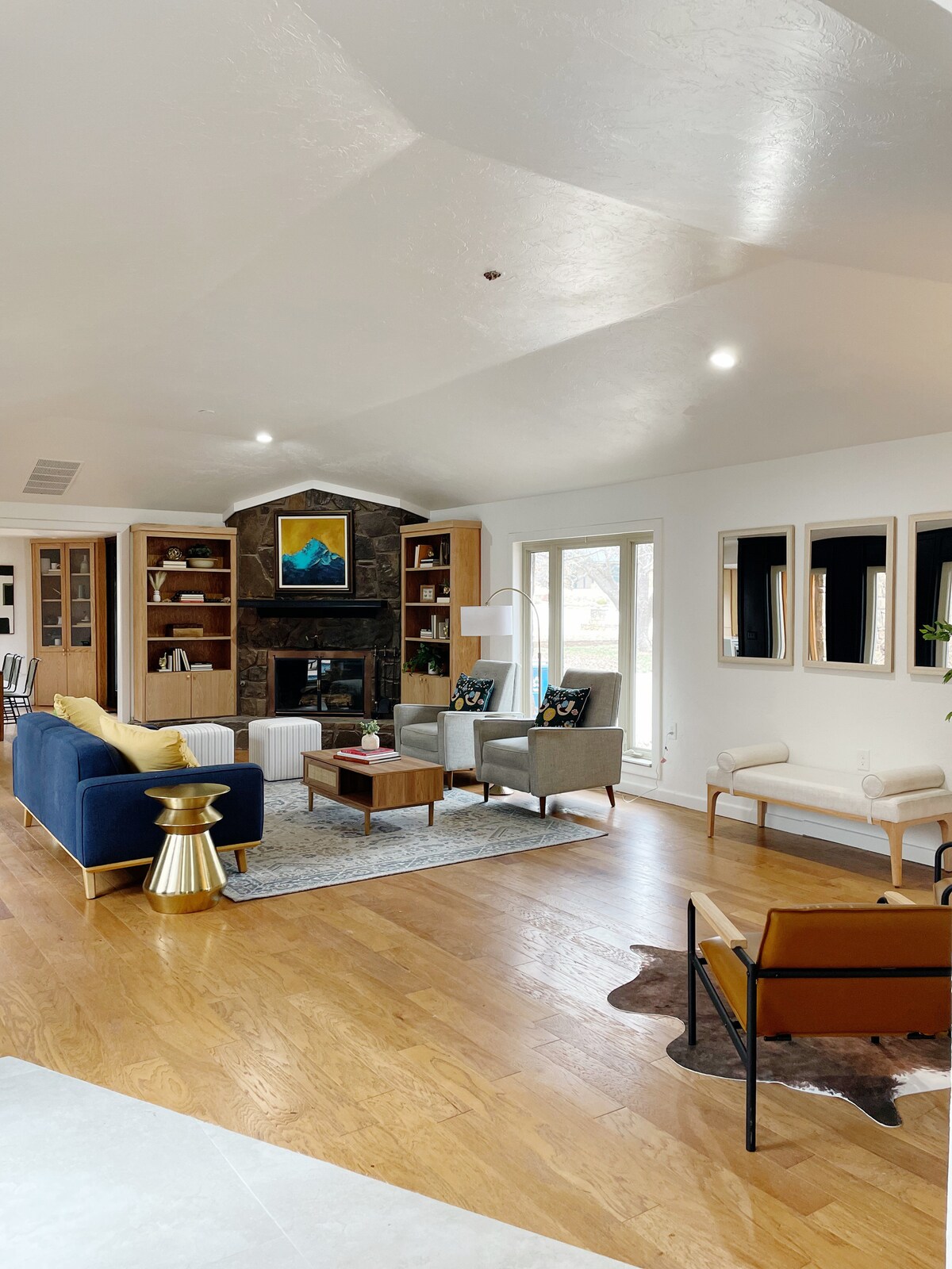 The living room showcases a mix of comfort and style, featuring a large sectional sofa, a coffee table, and various seating options. Natural light floods the space through multiple windows, highlighting the artful decor, defined by a stone fireplace and wooden details.