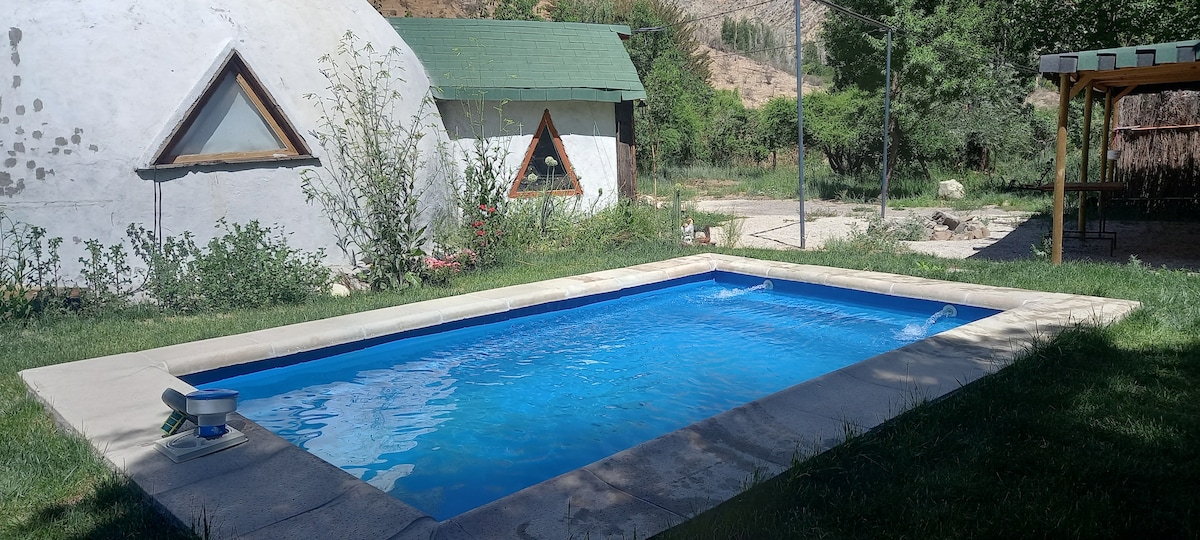 A private swimming pool is surrounded by well-kept grass and vegetation. Two structures with triangular windows can be seen in the background, with a shaded area nearby, providing a peaceful setting for relaxation.