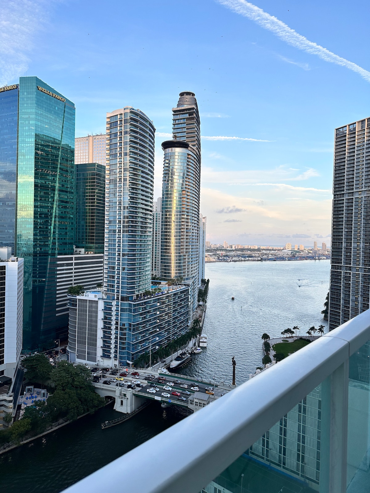 A scenic view from a 17th-floor balcony showcases the skyline of Brickell, featuring a blend of modern high-rise buildings. The river below reflects the sky, while the horizon displays a gentle transition of colors in the evening light.