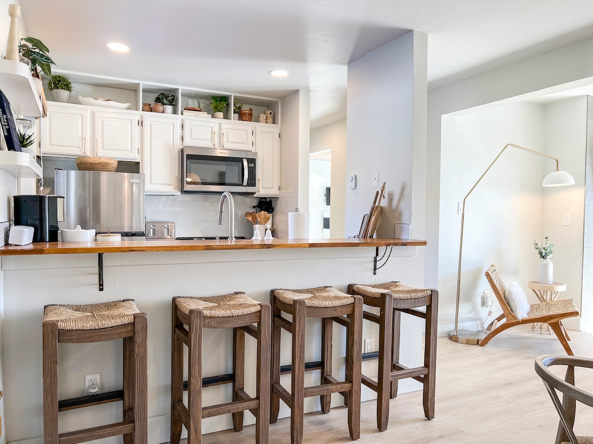 The kitchen features modern appliances, including a microwave and coffee maker, with wooden bar stools positioned at the counter. Natural light brightens the open space, which connects to a cozy sitting area on the right.