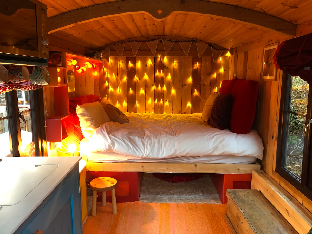 The interior of the shepherd's hut features a spacious bed with white linens, surrounded by warm string lights that illuminate the wooden walls. A colorful rug is placed on the floor, and a small stool is positioned near the kitchen area, creating a welcoming and cozy atmosphere.