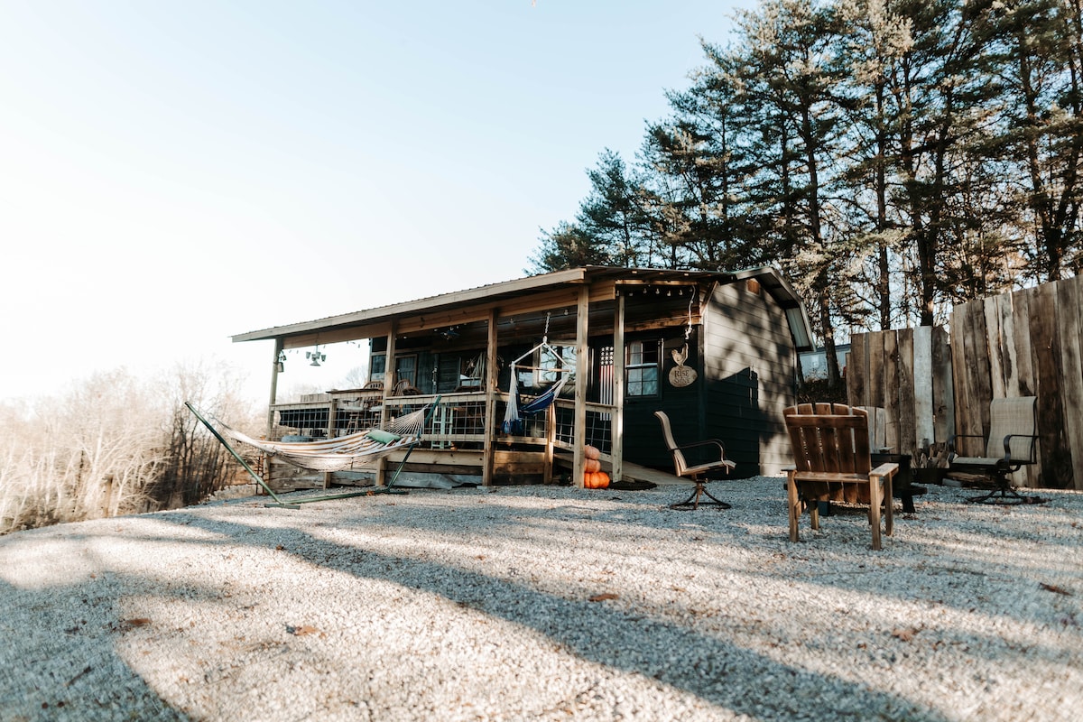 A cozy tiny cabin is set amid a gravel area, surrounded by trees. A hammock is gently draped between posts, while wooden chairs are placed nearby. The cabin features a porch with a railing and string lights, extending a welcoming atmosphere.