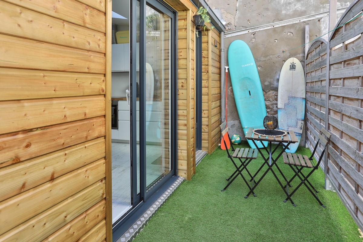 An exterior view of a small terrace is shown, featuring artificial grass and a wooden wall. A round table with two folding chairs is set up, accompanied by surfboards leaning against the wall, creating a relaxed outdoor space.