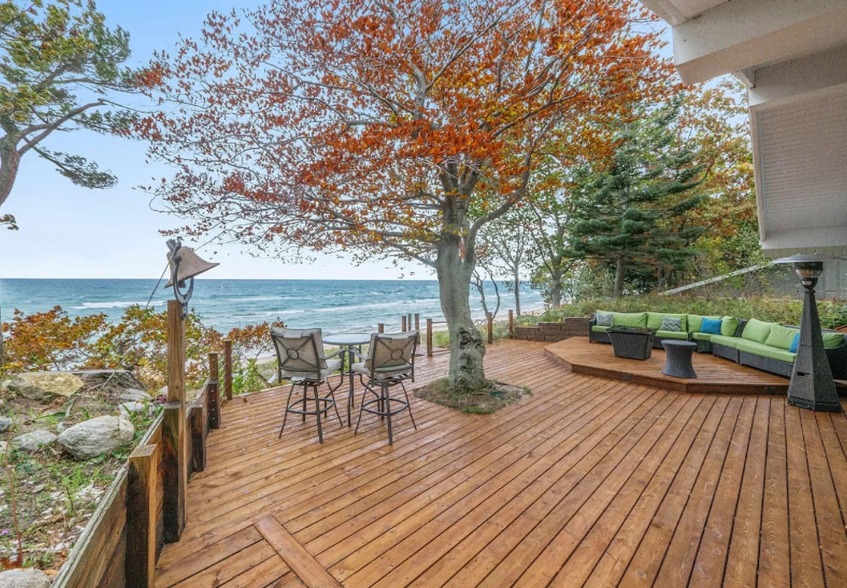 A spacious wooden deck extends towards Lake Michigan, bordered by colorful trees. Two seating areas are visible, showcasing a circular table with four chairs and a cozy sectional with green cushions. The serene lake water glimmers in the background, framed by lush foliage.