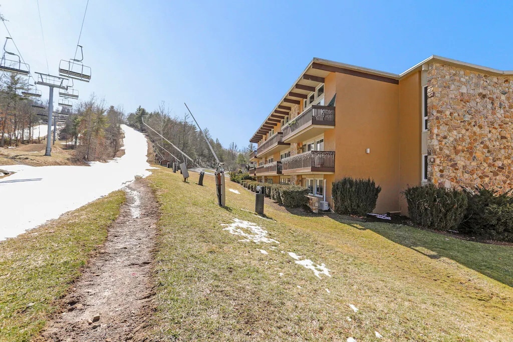 The exterior view of the condo shows a three-story building, featuring balconies with dark railings. A grassy area leads up to ski slopes, which are partially covered with snow. The sky is clear and blue, complementing the natural surroundings.
