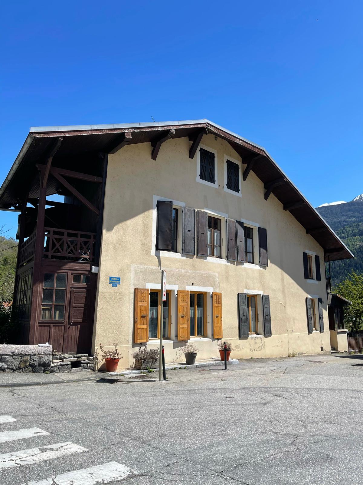 The exterior of a spacious mountain house is highlighted under clear blue skies. Traditional wooden shutters frame the windows, and a large balcony extends from the upper level. A well-maintained garden area with potted plants is visible at the entrance.