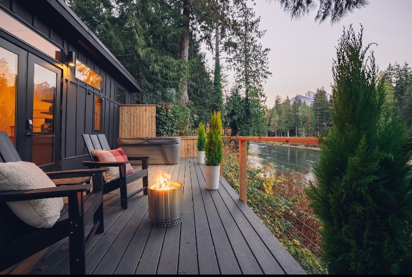 A serene outdoor deck is adorned with comfortable seating and a modern fire pit, surrounded by lush greenery. The tranquil river is visible beyond the railing, complemented by scenic mountain views in the background, creating a welcoming environment for relaxation.