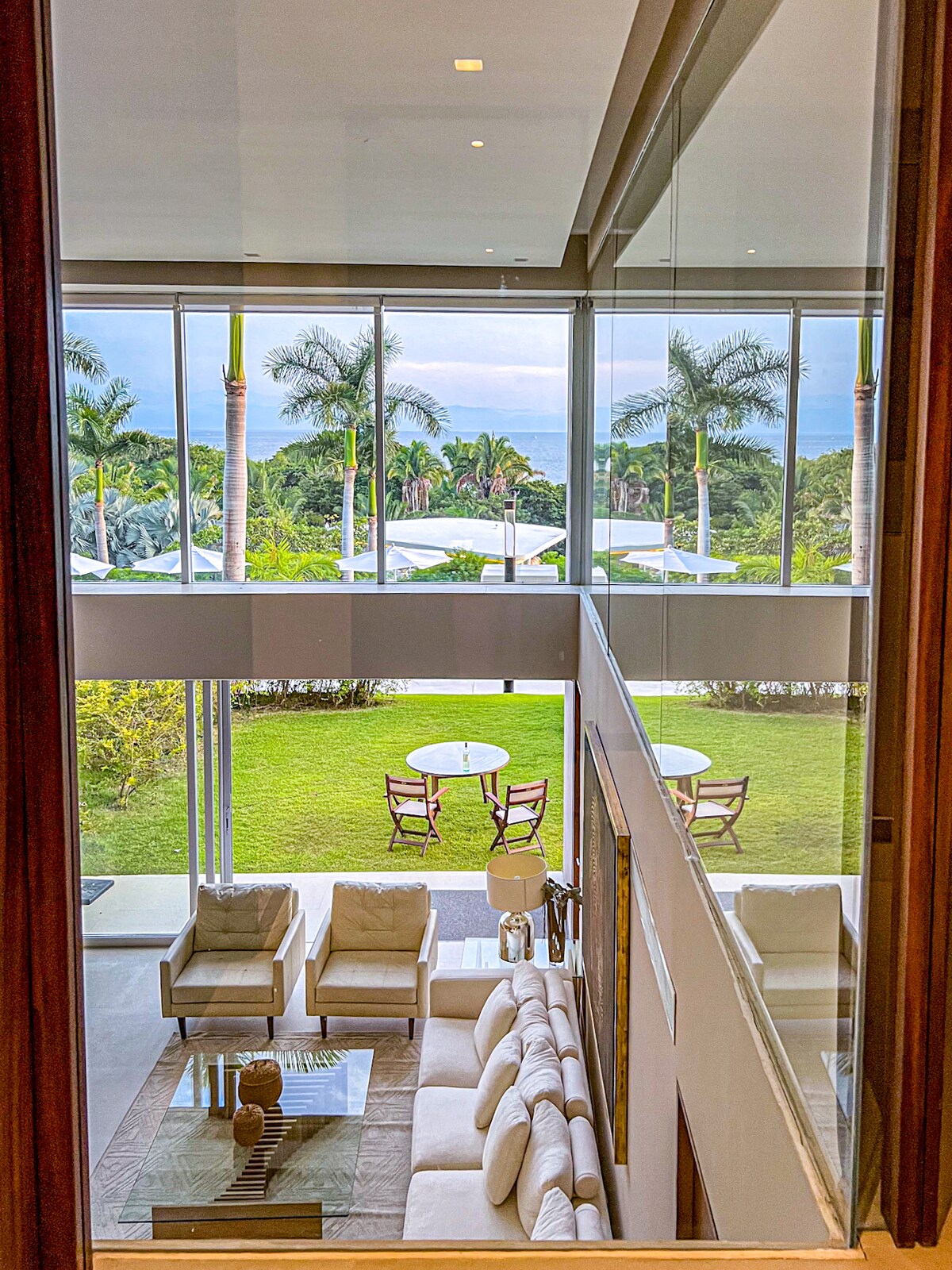 A spacious living area is visible through large glass windows. The room features a light-colored sofa facing a coffee table. Lush greenery and ocean views are seen from the garden outside, where outdoor seating is arranged on a patio.
