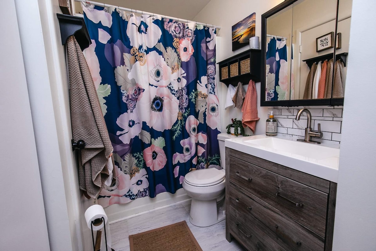 A bathroom features a floral shower curtain in soft pastel colors complemented by a sleek white countertop. A wooden vanity offers storage beneath a wall mirror, while towels hang neatly on hooks. A small rug is placed in front of the toilet for added comfort.