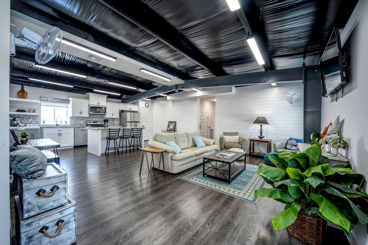 The open concept living area features a spacious seating arrangement with a light-colored sofa, accompanied by a coffee table and a decorative area rug. The kitchen area is visible, showcasing modern appliances and bar seating. Natural light illuminates the room, highlighting indoor plants and decorative elements.
