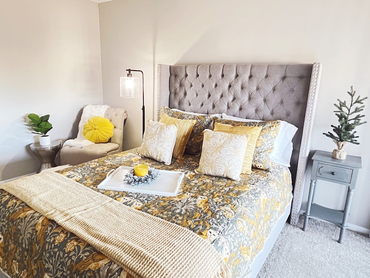 A comfortable bedroom features a tufted gray headboard with decorative pillows in yellow and floral designs. A side table holds a small plant and a yellow decorative object. A cozy chair is positioned nearby, creating a restful corner, with soft natural light illuminating the space.