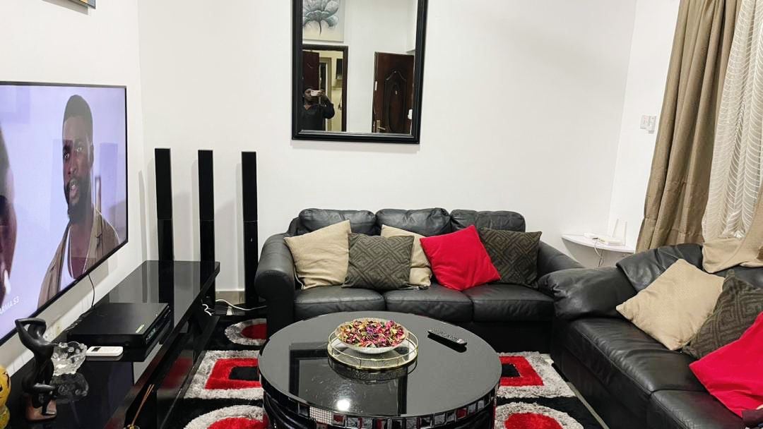 A modern living room features a large black leather sectional sofa adorned with red and gray pillows. A circular black coffee table sits in the center, holding a decorative tray of colorful items. A flat-screen TV and tall speakers are positioned nearby, with ambient lighting illuminating the space.