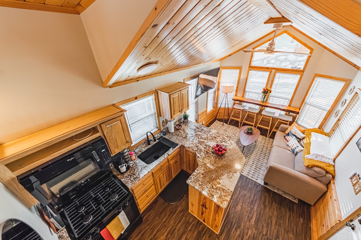 An overhead view showcases the open-concept living area with a high, wooden ceiling. The fully equipped kitchen features a granite countertop and modern appliances. A cozy seating area with large windows allows natural light, while a designated workspace is highlighted near the back, ideal for remote work.