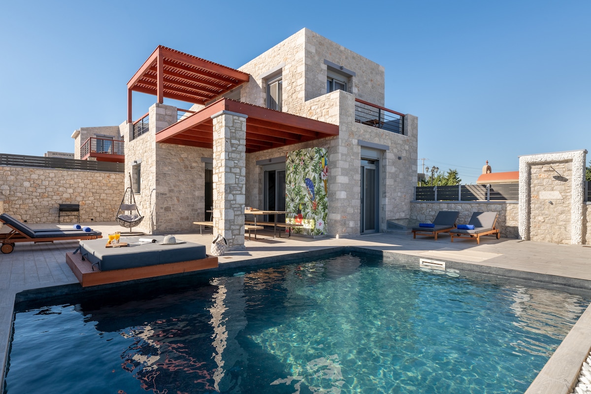 A modern villa is shown with two levels, featuring a stone facade and a red roof. A swimming pool sits in the foreground, with lounge chairs arranged nearby. An outdoor dining area and a decorative mural add character to the space, surrounded by a private courtyard.