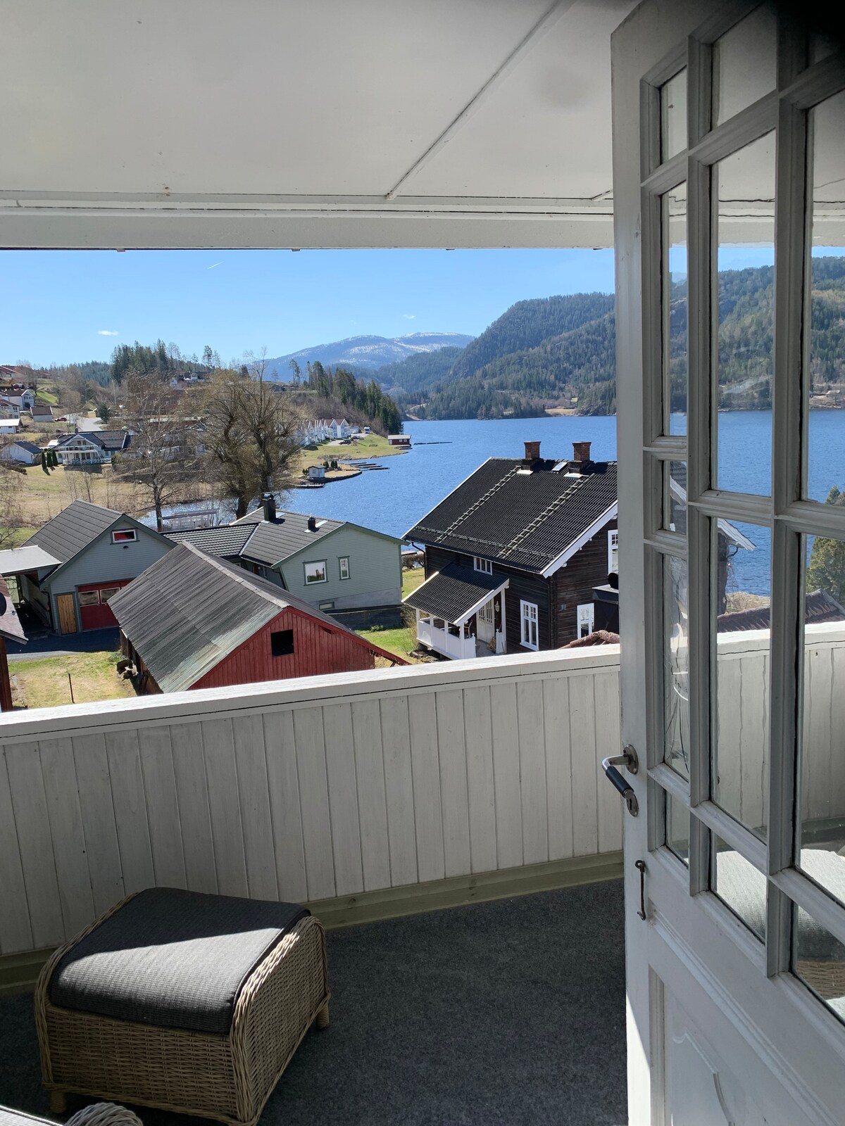 A scenic view of the fjord opens through a doorway adorned with glass panels. The charming village is visible, featuring a mix of colorful buildings along the waterfront. Lush mountains rise in the background, completing this tranquil landscape.