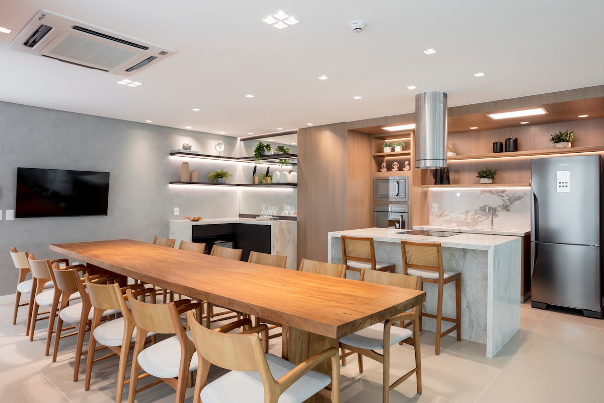 A spacious dining area features a large wooden table surrounded by light wooden chairs. The open kitchen showcases modern appliances and minimalist cabinetry, while under-cabinet lighting enhances the sleek design. A television is mounted on a wall, complementing the overall contemporary aesthetic.