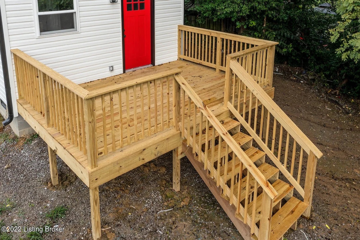 An accessible wooden deck is featured, complete with a wide ramp and stairs. The railing is designed for safety, and the deck connects to a white exterior with a bright red door, surrounded by greenery.