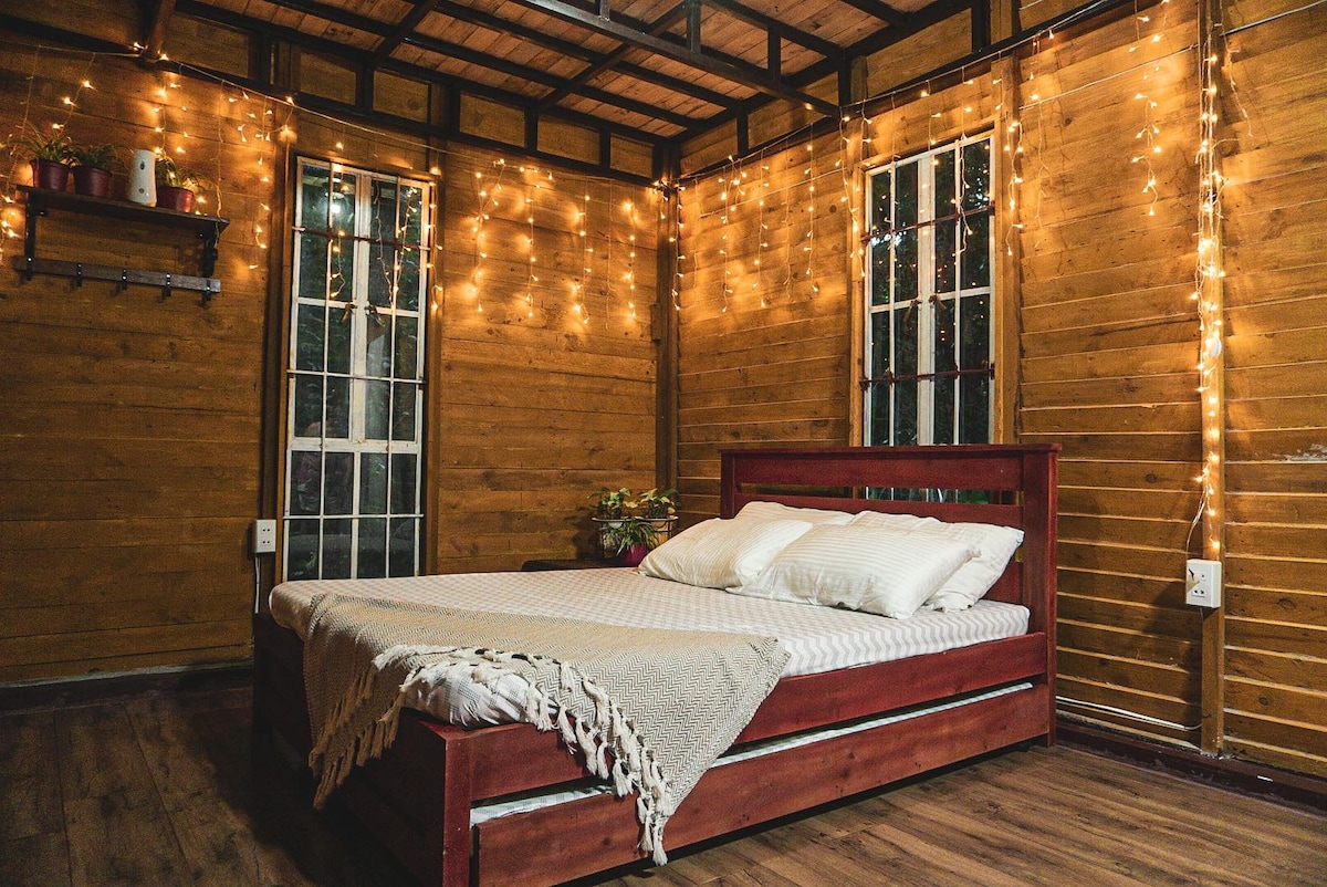 A cozy bedroom is highlighted by warm wooden walls and soft lighting created by string lights. A spacious bed sits prominently in the center, adorned with neutral-toned linens. Windows allow for natural light, enhancing the inviting feel of the room.