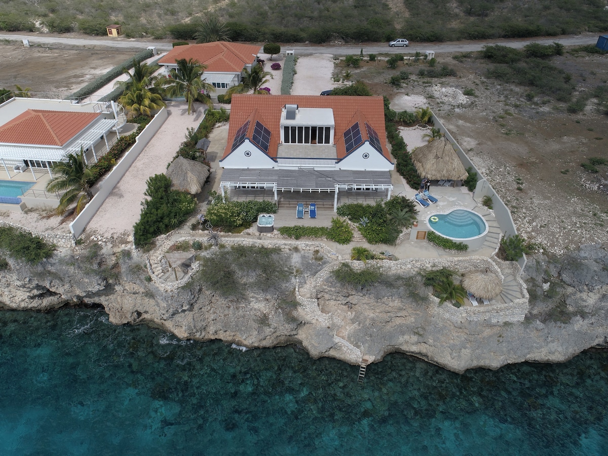 An aerial view of a villa positioned on a cliff overlooking the Caribbean Sea. The property features a red-tiled roof, solar panels, and a blue swimming pool surrounded by tropical gardens. Direct access to the sea is visible, along with a staircase leading down to the water.