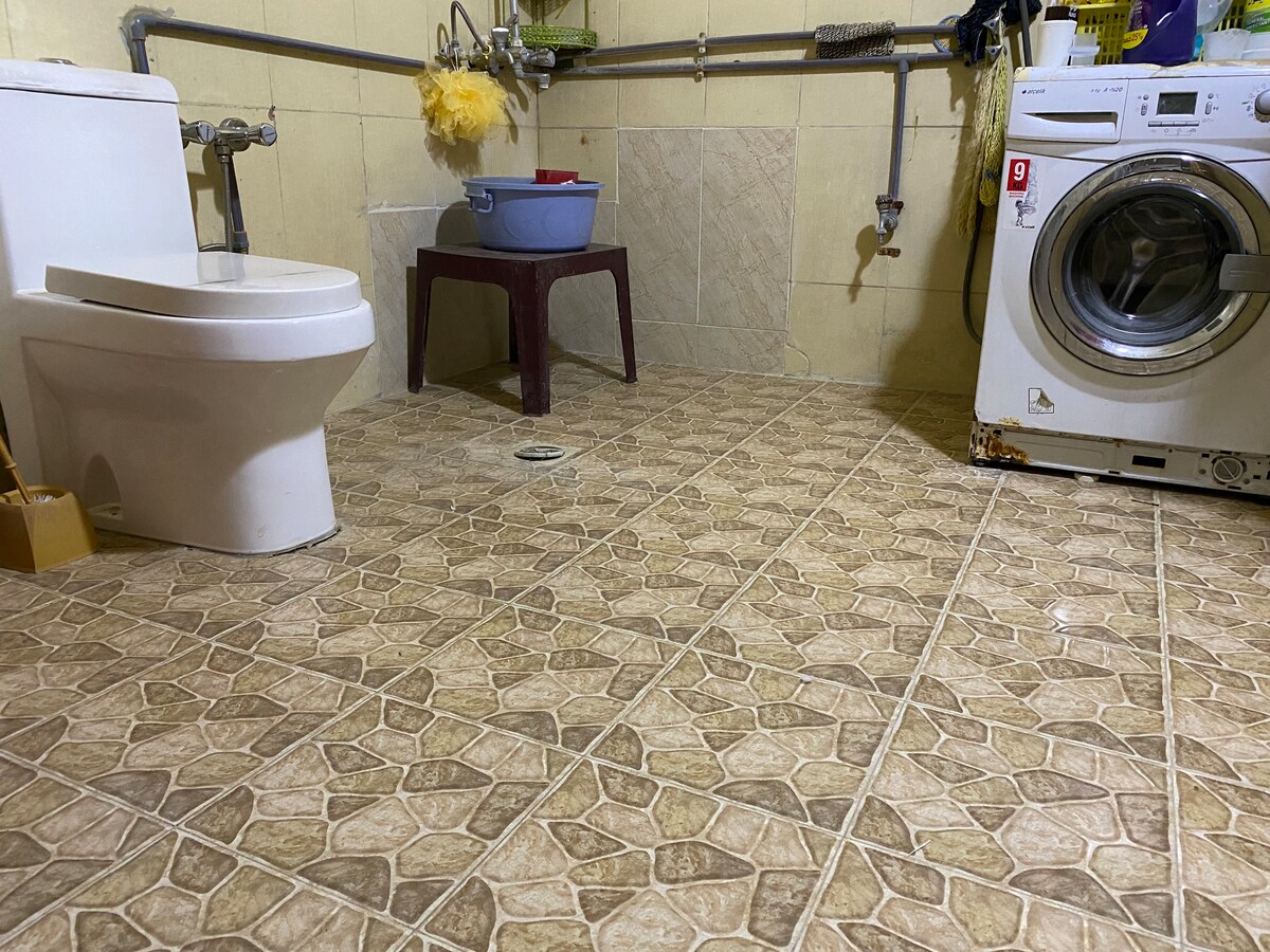 A clean bathroom is depicted, featuring a white toilet and a washing machine beside it. A plastic stool is placed nearby, while a blue basin rests on the stool. The tiled floor displays a textured pattern, and the walls are painted with a muted hue.
