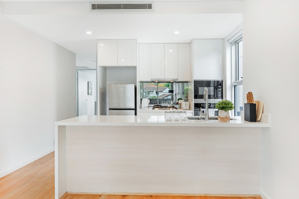 The modern kitchen showcases sleek white cabinets and a spacious island counter. Bright natural light filters through large windows, illuminating premium appliances, including a refrigerator and oven. The kitchen is well-equipped with essential utensils, inviting cooking and dining experiences.