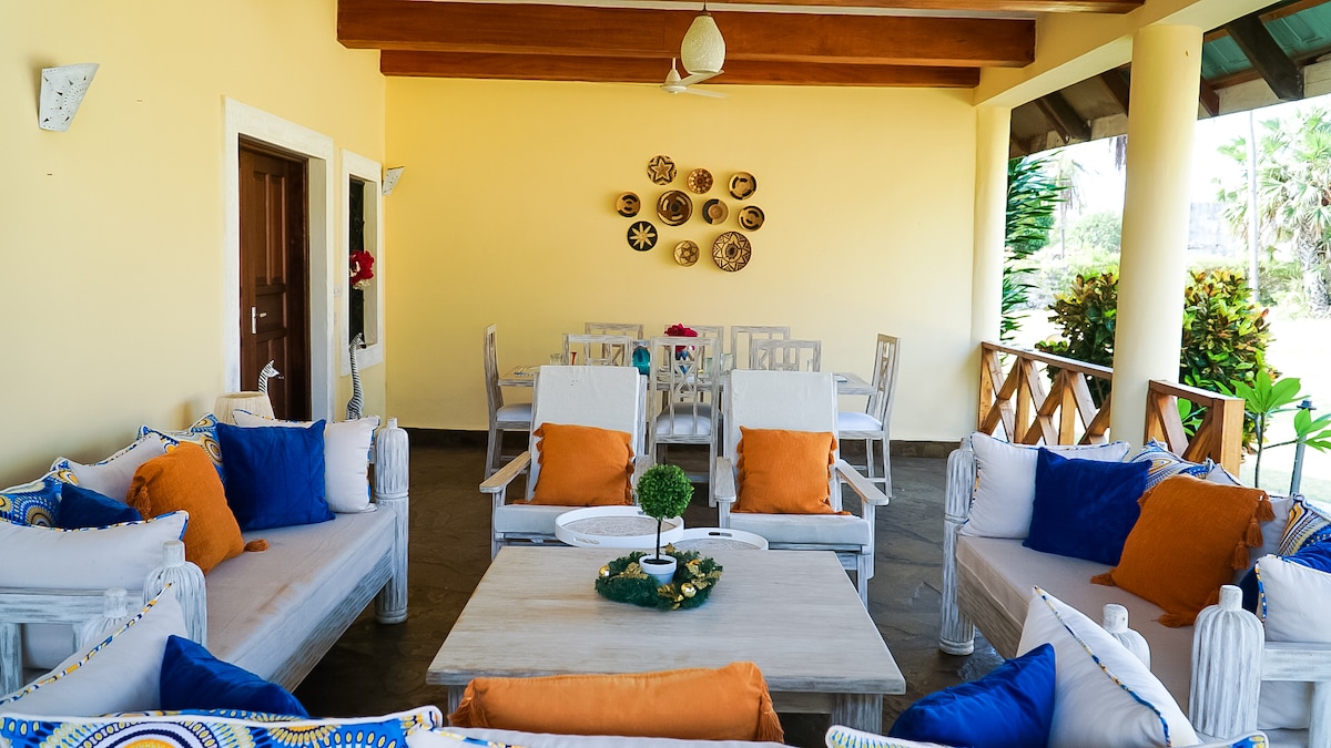 An outdoor seating area features comfortable couches adorned with vibrant blue and orange cushions. A wooden table sits at the center, surrounded by a dining table set for six. The space is bathed in natural light, with decorative wall plates adding a touch of charm.
