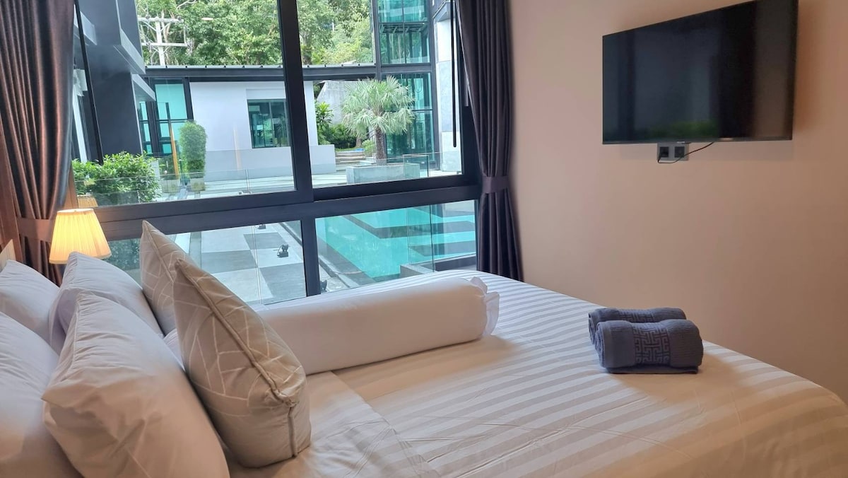 A well-appointed bedroom features a spacious bed adorned with decorative pillows, offering a view of the outdoor pool through large glass windows. A cozy blanket is neatly arranged alongside a rolled towel, while soft natural light fills the room.