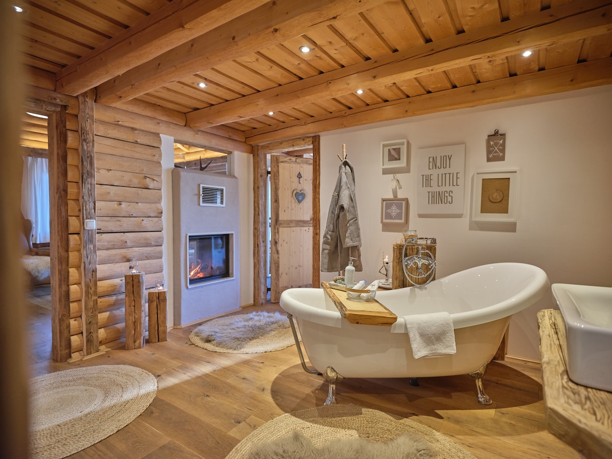 The bathroom features a freestanding bathtub accompanied by a wooden tray. Soft lighting highlights the warm wooden ceiling and walls. A cozy rug lies underfoot, and the space is complemented by decorative wall art and a fireplace, creating a serene atmosphere.