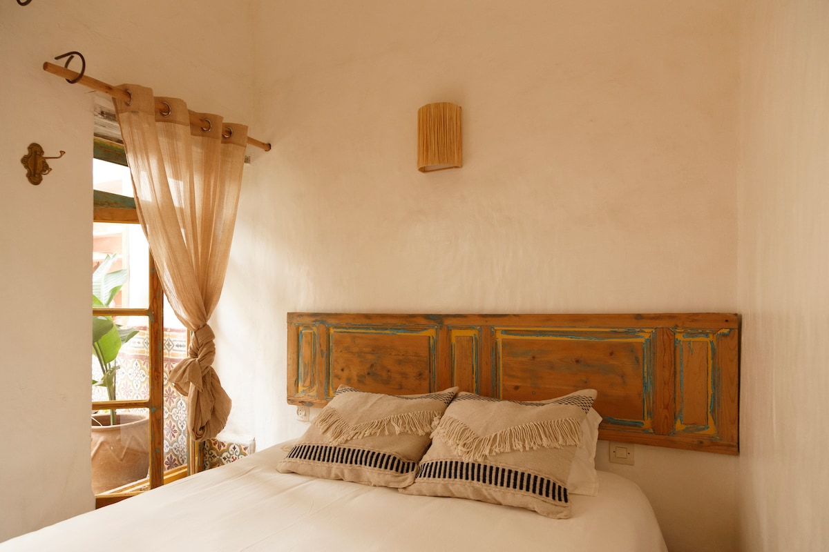 A bright bedroom features a king-sized bed, adorned with two textured pillows. A wooden headboard adds rustic charm to the space. Natural light filters through sheer curtains, illuminating the room and revealing a small potted plant visible through the window.