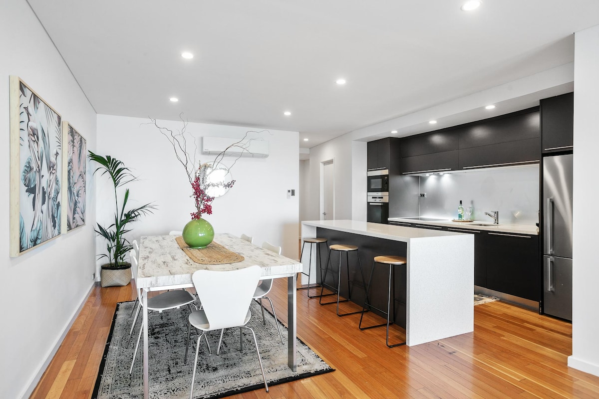 An open-plan kitchen and dining area is displayed, featuring a spacious dining table with decorative elements. Modern kitchen cabinets, appliances, and a breakfast bar are visible, complemented by warm wooden flooring and soft lighting that enhances the contemporary design.