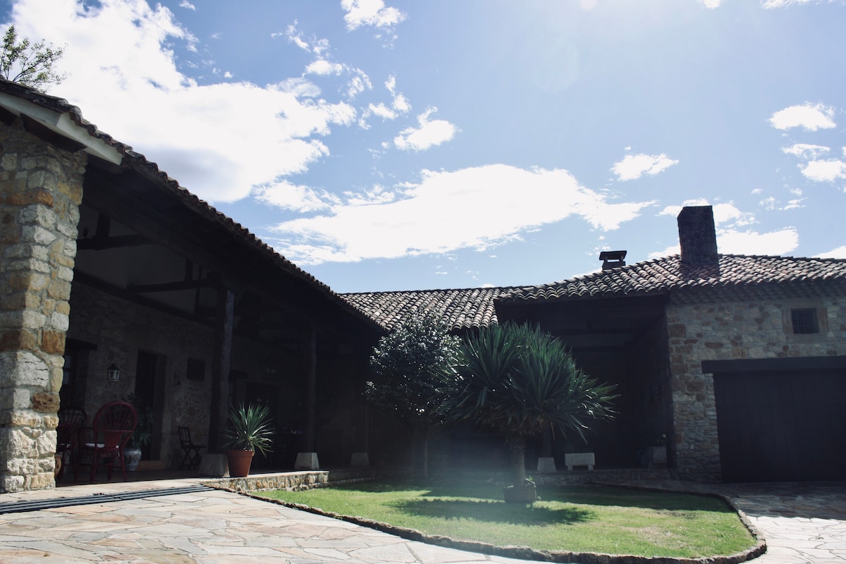 A charming exterior view features a stone facade, traditional roof tiles, and lush greenery. Sunlight brightens the space, highlighting a spacious patio area with stone flooring. The entrance is flanked by potted plants, welcoming guests to the home.