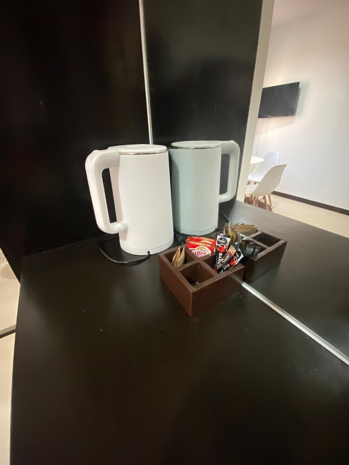 Two electric kettles, one white and one light blue, are positioned on a dark surface. A wooden caddy containing various tea and coffee pods is nestled between them, providing convenient access to beverages.