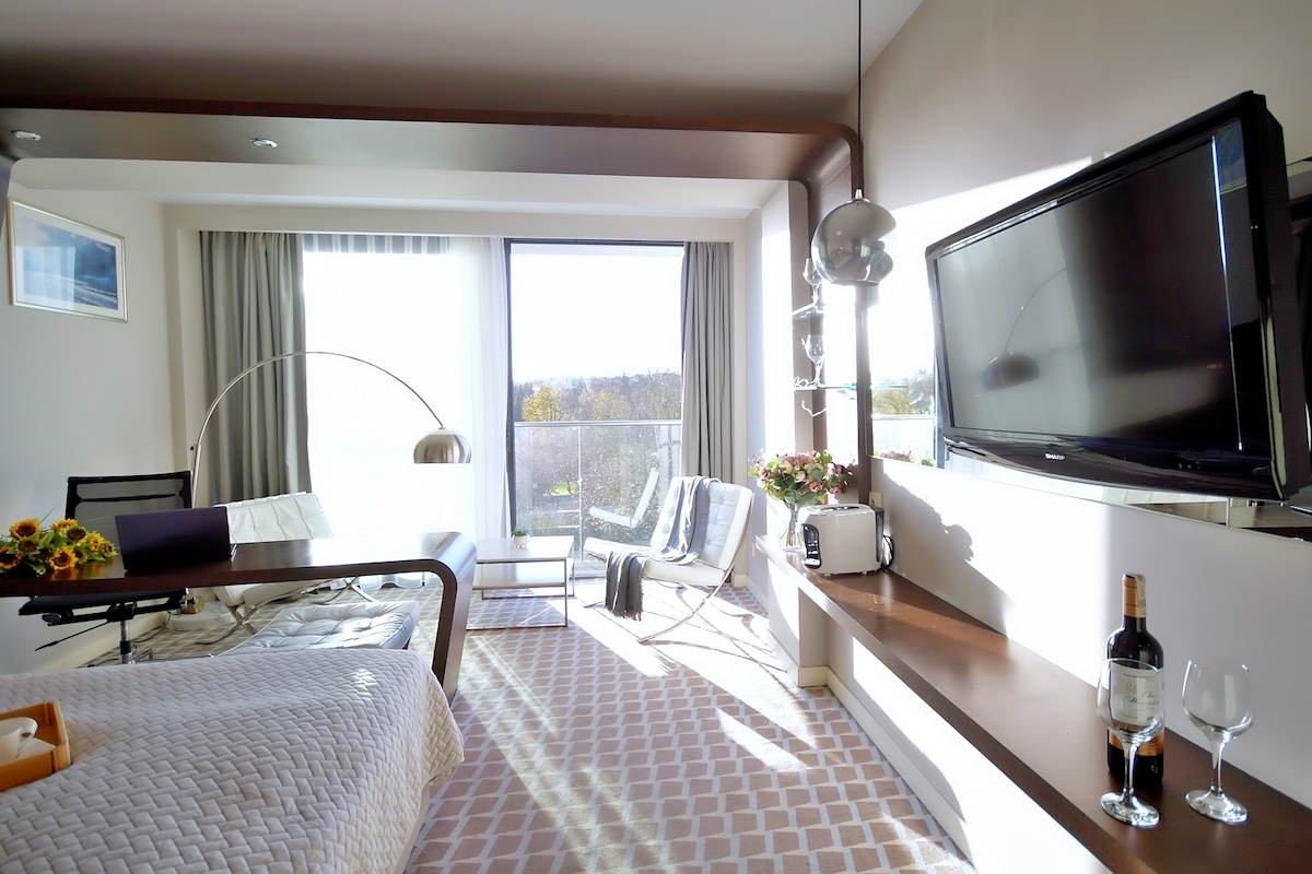 A modern room features large glass doors that open to a scenic view. Sunlight fills the space, highlighting a spacious bed with a textured blanket. A flat-screen TV is mounted on the wall, and a small dining area with wine bottles and glasses is visible.