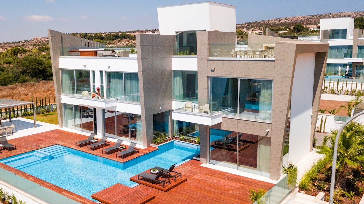 A modern villa is displayed, featuring large glass walls that offer a view of the inviting outdoor pool and surrounding greenery. A spacious wooden deck with lounge chairs is positioned beside the pool, creating a relaxed outdoor space.