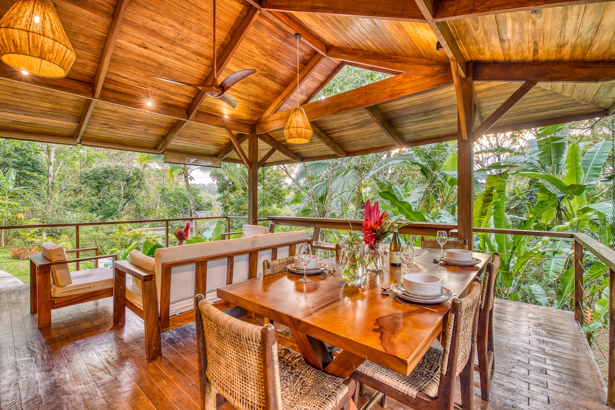 An open-air dining area showcases a wooden table set for several, complemented by wicker chairs. Natural light filters through a thatched ceiling, highlighting the surrounding greenery. A cozy seating arrangement with light-colored cushions offers a relaxed space adjacent to the dining setup.