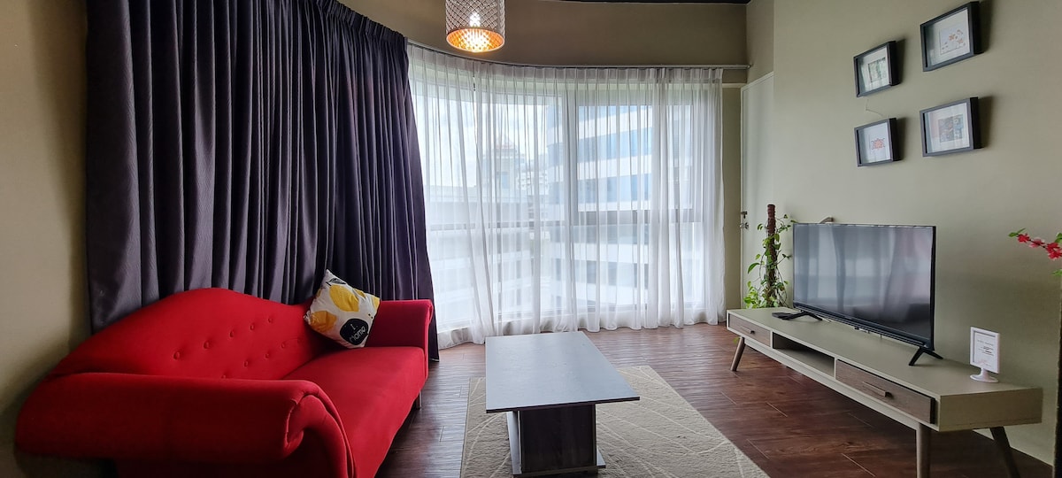 A bright living space features a red sofa paired with a decorative pillow, positioned alongside a light-colored coffee table. Large windows are dressed in sheer curtains, allowing natural light to fill the room. A television sits on a low console against a neutral wall.