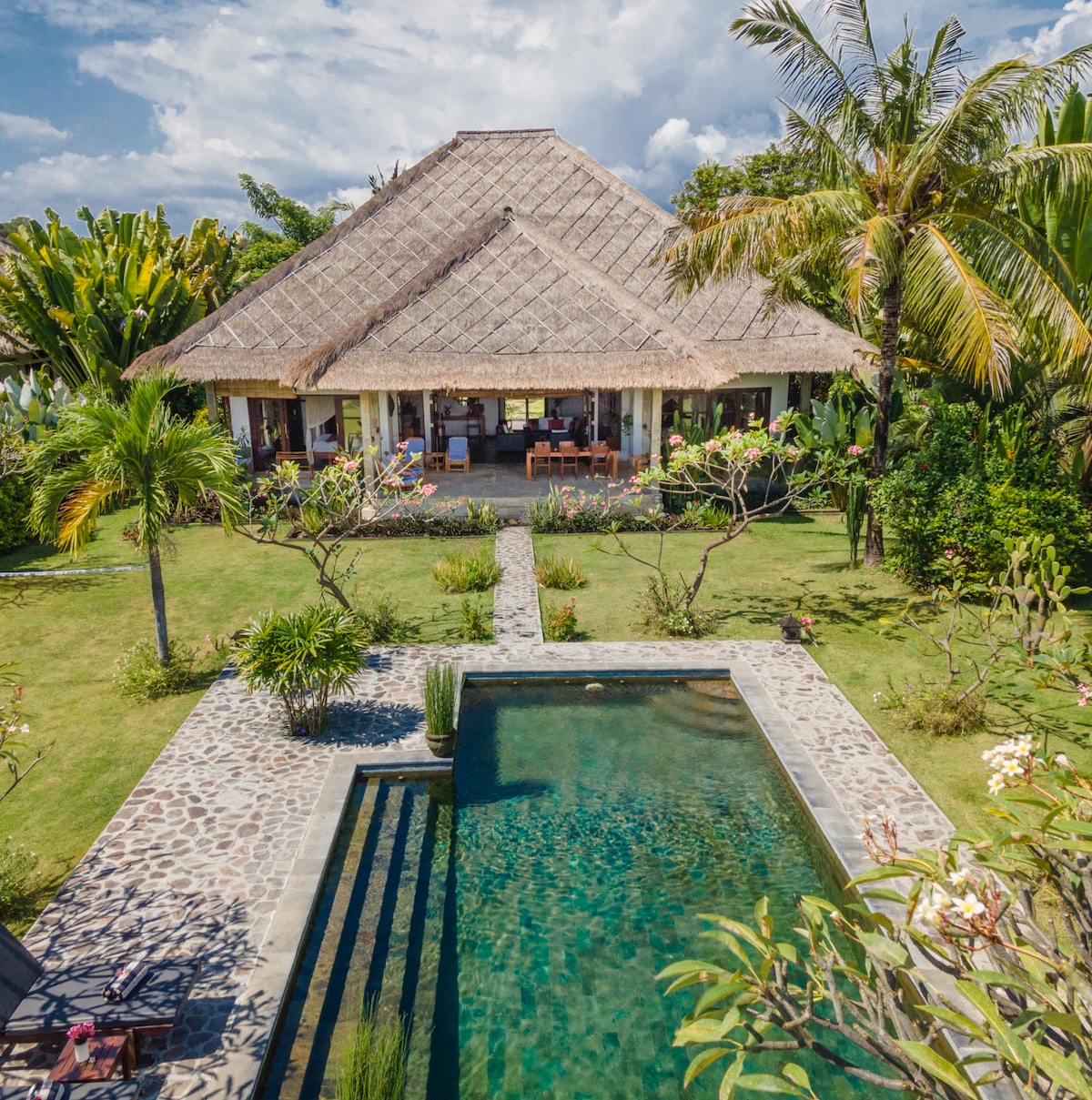 The villa features a spacious pool surrounded by lush tropical greenery, with a stone pathway leading to the entrance. The traditional thatched roof complements the serene environment, while the open design showcases inviting indoor dining and lounging areas.
