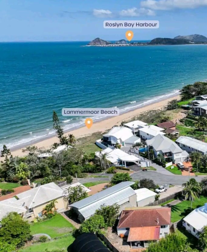 Aerial view captures the coastal landscape, showcasing sandy Lammermoor Beach lined by residential buildings. The sparkling water of the harbor glistens in daylight, with distant shores visible in the background. Lush greenery frames the scene, hinting at a serene beachside community.