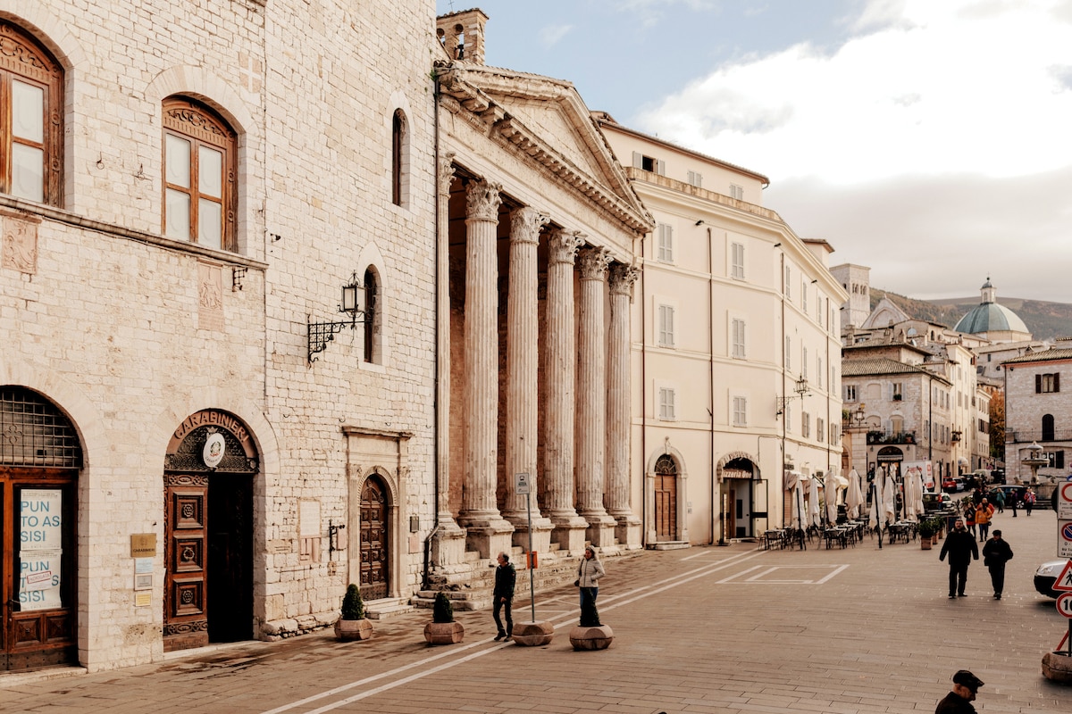 The image captures a historic square in Assisi, featuring grand columns of a classical building alongside charming stone facades. People stroll on the stone-paved street, with diverse shops and cafés visible in the background, offering a lively ambiance in the heart of the city.