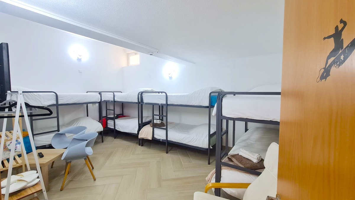 A shared room features four sets of bunk beds, accommodating eight individual sleeping spaces. A small table and a chair are visible, providing seating. Soft lighting is provided by wall-mounted fixtures, while a door entry is seen on the right side.