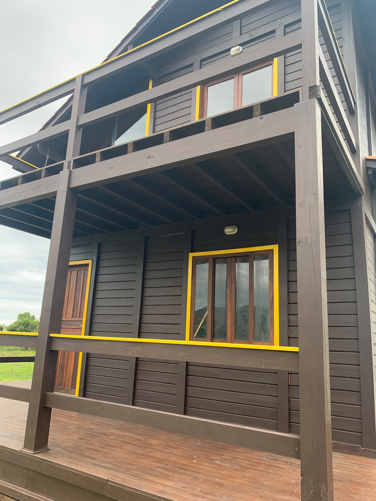 The two-story cabin features a dark wood exterior with vibrant yellow trim around the windows and doors. A spacious wooden deck extends in front, providing an inviting entryway. Large windows enhance natural light and offer views of the surrounding landscape.