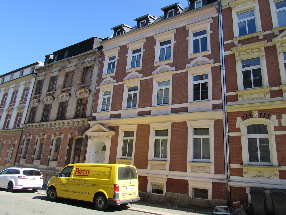 A historical building features an elegant facade with a combination of red brick and cream-colored detailing. Large windows are symmetrically arranged across three stories, allowing natural light to fill the interior. A yellow van is parked in front, contrasting the muted tones of the structure.