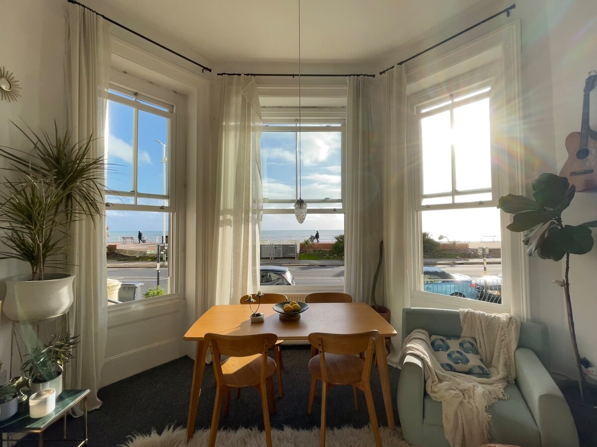 A bright dining area features a wooden table surrounded by four chairs. Large windows allow natural light to fill the space, offering views of the sea. Light curtains gently frame the windows, and a comfortable sofa is positioned nearby, creating a welcoming atmosphere.