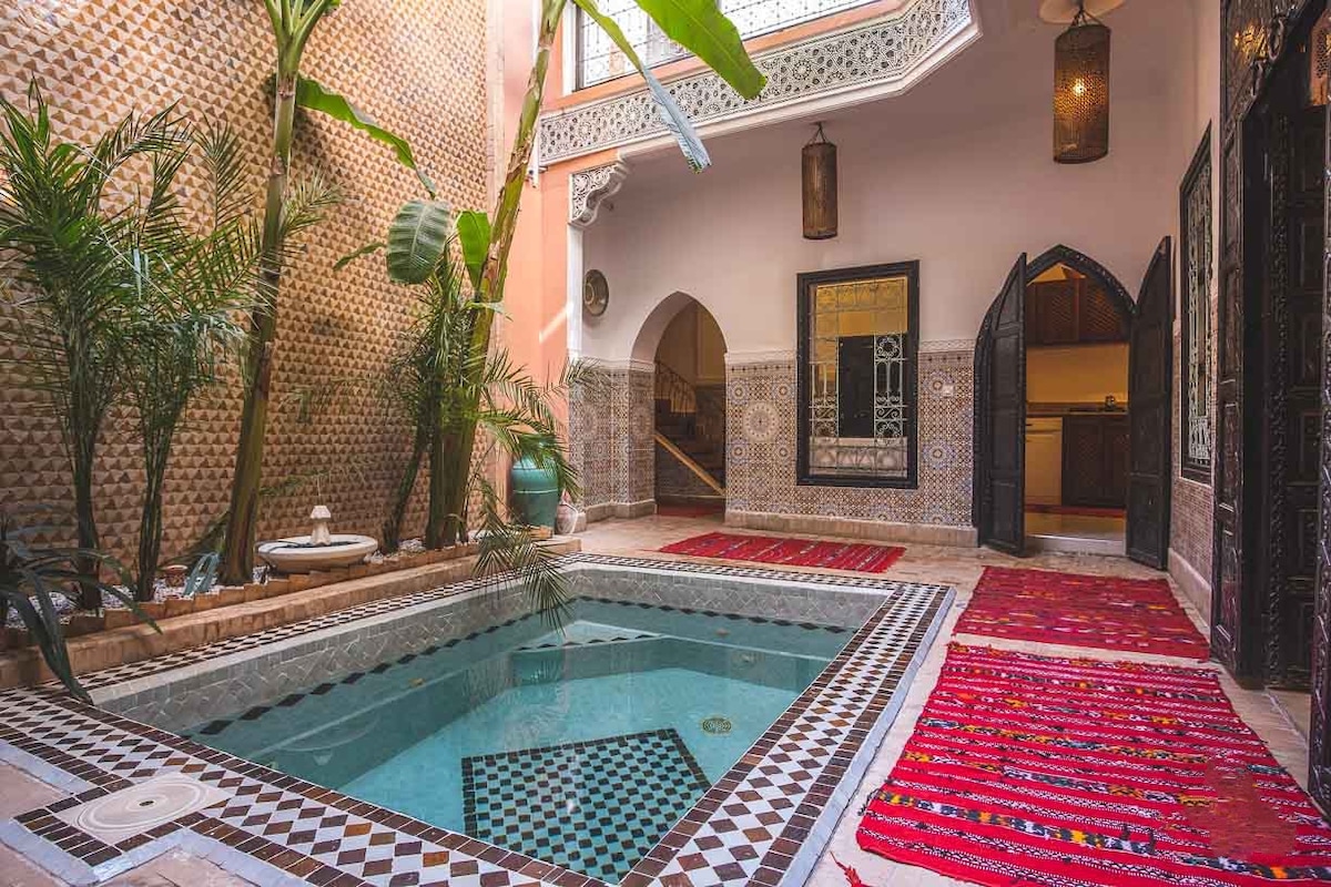 A serene patio area is highlighted by a small swimming pool, surrounded by decorative tile work and lush green plants. Colorful rugs are laid across the floor, and arched doorways lead to additional spaces, creating a harmonious indoor-outdoor atmosphere.