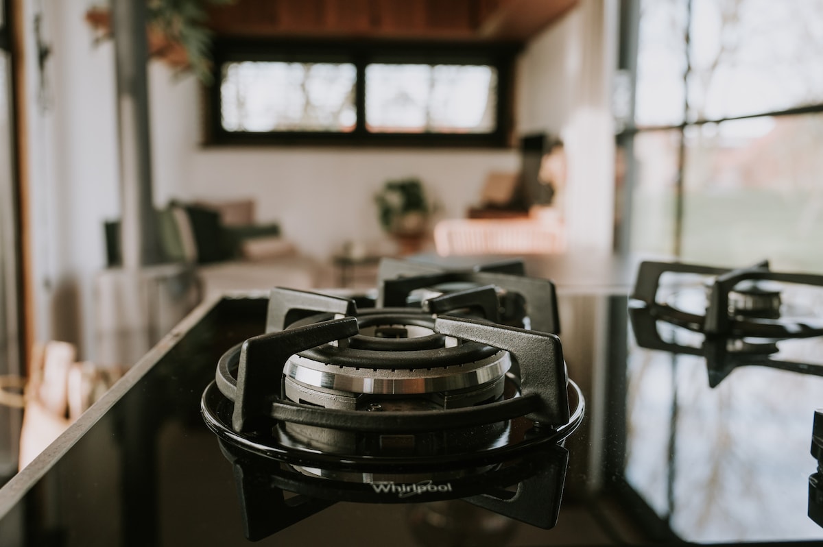 A modern gas stove is prominently featured in the kitchen area, showcasing sleek black burners and a polished surface. In the background, natural light filters through large windows, illuminating a cozy seating area with minimalist decor.