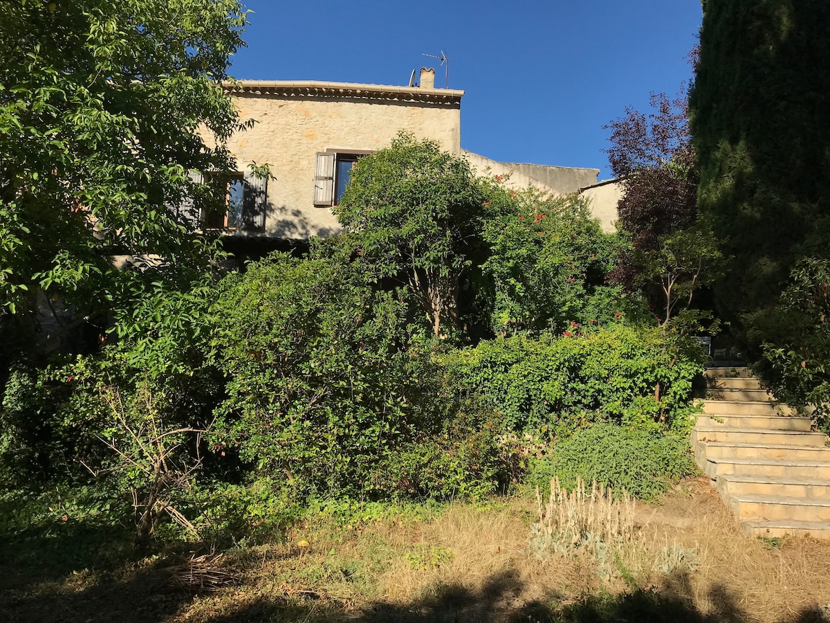 The exterior of a charming stone house is surrounded by lush greenery, featuring various plants and trees. A set of stone steps leads to the entrance, framed by the vibrant foliage that enhances the tranquil garden setting.