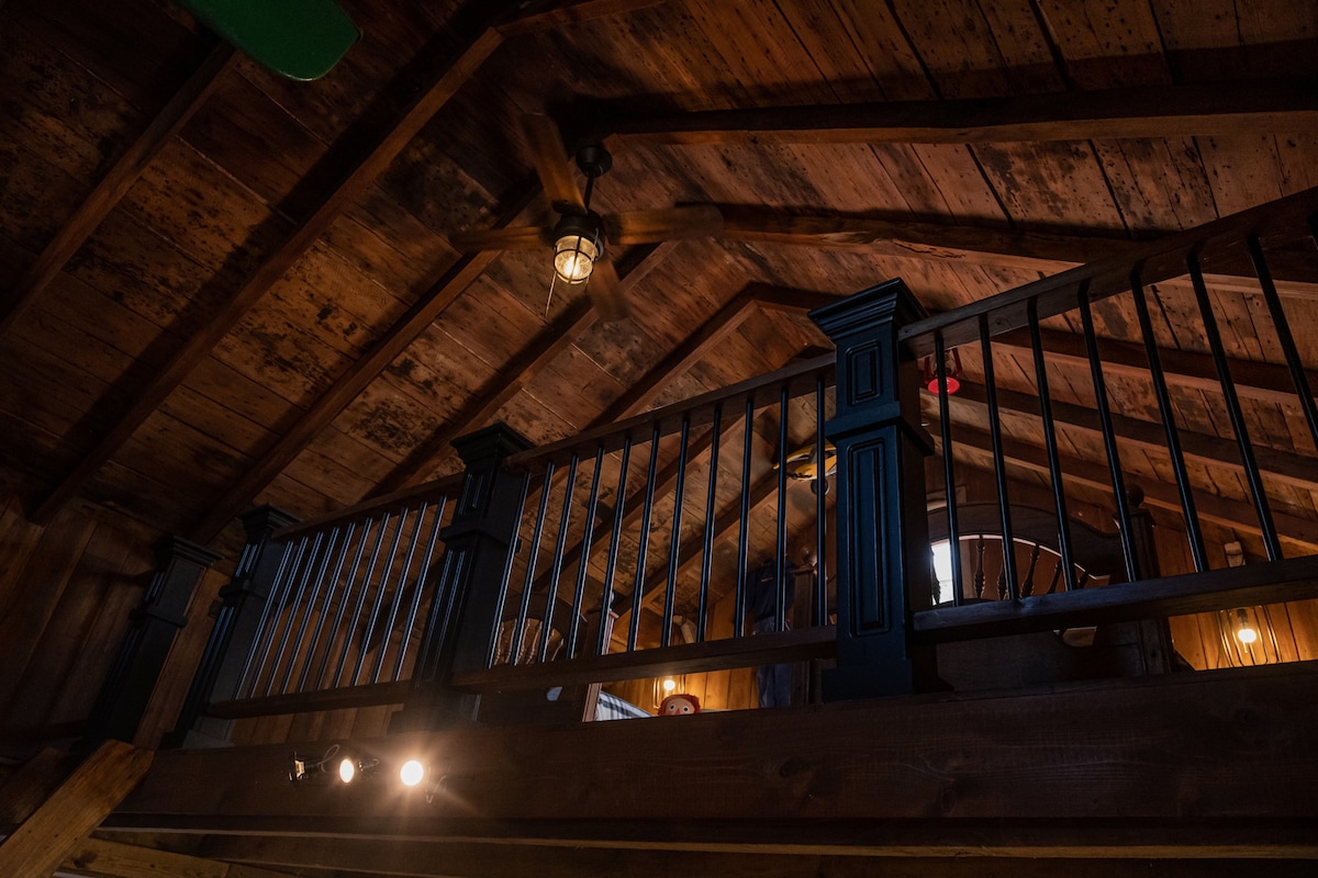 A view from below showcases a wooden loft, featuring a railing with vertical metal balusters. The ceiling is adorned with exposed beams, highlighting the rustic charm of the cabin. Soft lighting emanates from a fixture, illuminating the warm tones of the wood.