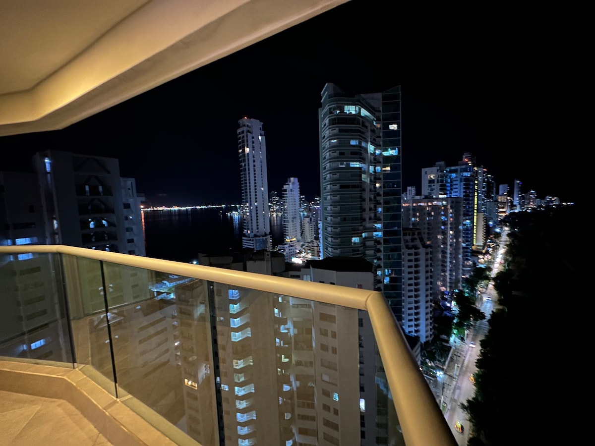 A night view from a modern balcony showcases the illuminated skyline of Cartagena, with tall buildings reflecting light along the water. The dark sky contrasts against the vibrant city lights, providing a captivating atmosphere of urban life.